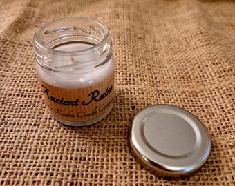 May include: A small glass jar candle with a white label that reads "Ancient Ruins" and "The Realm Candle Company". The candle is sitting on a brown burlap surface. The candle has a silver lid.