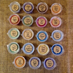 May include: An overhead shot of 18 small, round wax melt containers with various colors and labels. The labels include names like "Ancient Ruins," "Dragon's Blood," and "Starry Night." The containers are arranged on a textured, tan surface.