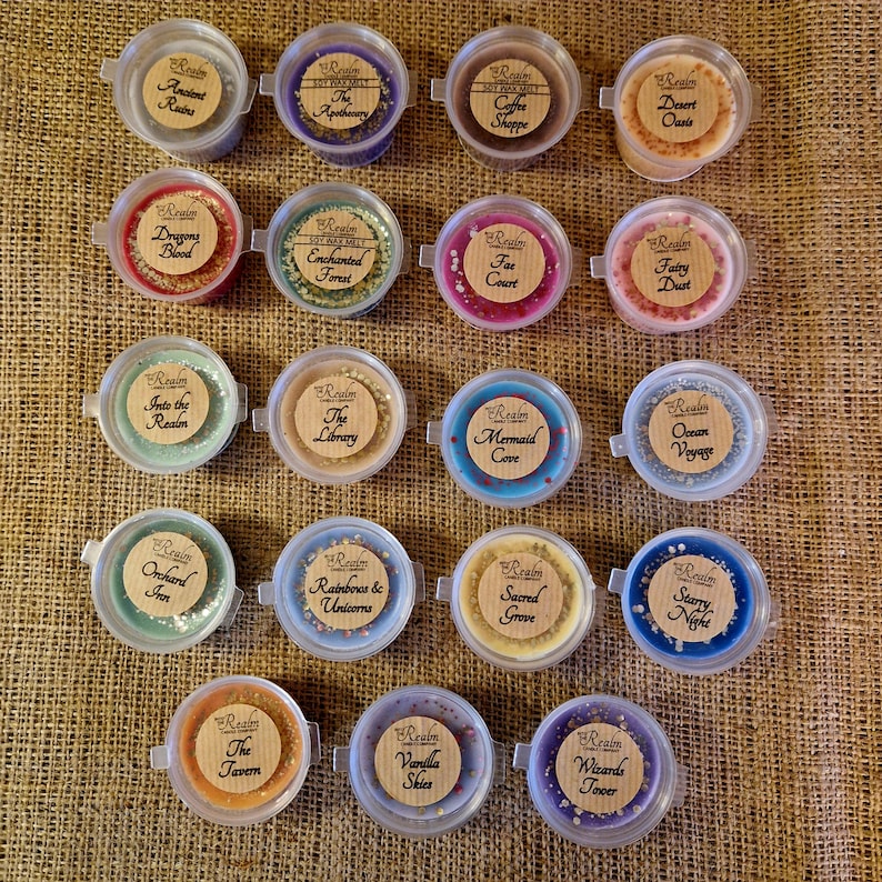 May include: An overhead shot of 18 small, round wax melt containers with various colors and labels. The labels include names like "Ancient Ruins," "Dragon's Blood," and "Starry Night." The containers are arranged on a textured, tan surface.