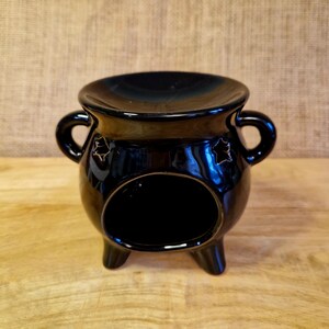 May include: A glossy black ceramic oil warmer with a round opening and star-shaped cutouts. The warmer has a small dish on top, two handles, and three small legs. The background is a light brown burlap.