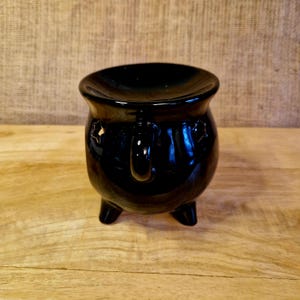 May include: A glossy black ceramic cauldron with three short legs and a handle. The cauldron has a wide opening and star-shaped cutouts on the sides. It sits on a light brown wooden surface.