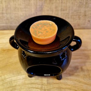 May include: A black ceramic wax warmer with a round dish holding an orange wax melt sprinkled with green herbs. The warmer has a cauldron shape with star cutouts and small feet. The background is a neutral tan.
