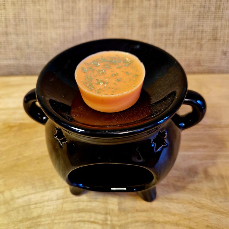 May include: A black ceramic wax warmer with a round dish holding an orange wax melt sprinkled with green herbs. The warmer has a cauldron shape with star cutouts and small feet. The background is a neutral tan.