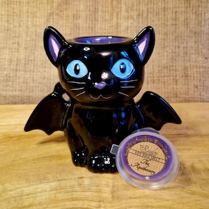 May include: A black ceramic cat-shaped candle holder with blue eyes, purple accents, and bat wings. A soy wax melt container with the text "The Apothecary" is next to the candle holder. The candle holder is on a wooden surface.