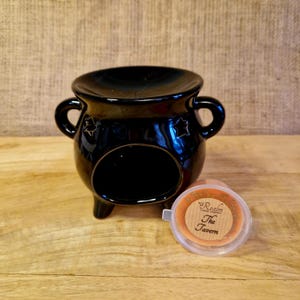 May include: Black ceramic wax melt warmer with a round top, two handles, and star cutouts. The warmer has three legs and a circular opening. A wax melt container with "The Tavern" is next to it.