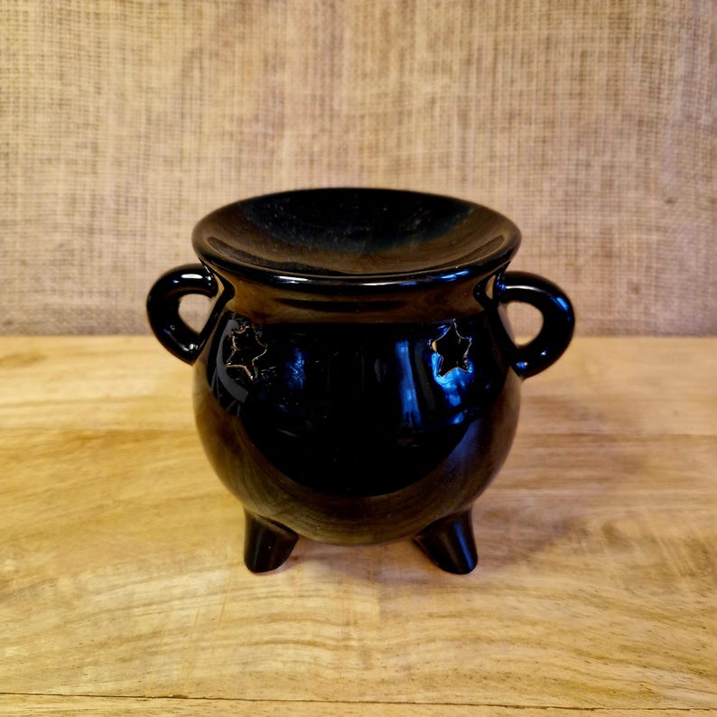May include: A glossy black ceramic cauldron with three legs and two handles. The cauldron has a wide rim and star-shaped cutouts on the sides. The cauldron is likely used for decorative or ritual purposes.