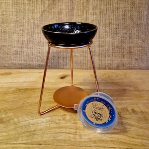 May include: A black and gold essential oil warmer with a copper-coloured metal stand. The bowl has a starry night design. The warmer is next to a stack of blue wax melts with the text "Starry Night".