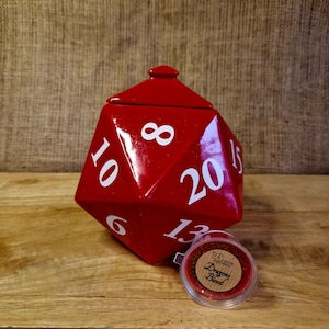 May include: A large, red, polyhedral die-shaped container with white numbers. A small, clear container with a label that reads "Realm Dragons Blood" is next to the die. The items are on a wooden surface.