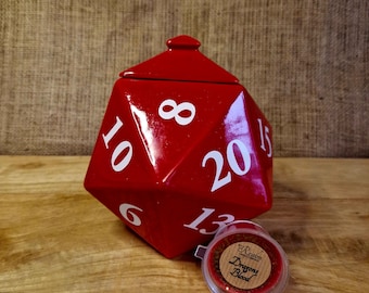May include: A large, red, polyhedral die-shaped container with white numbers. A small, clear container with a label that reads "Realm Dragons Blood" is next to the die. The items are on a wooden surface.