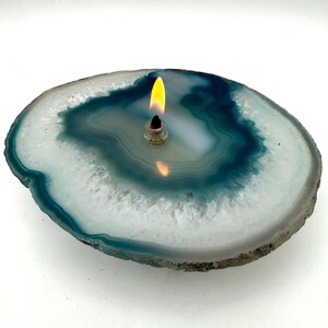 May include: A lit candle in a clear glass holder sits atop a polished agate slice. The agate features concentric bands of white, teal, and dark blue. The flame is bright yellow and orange.
