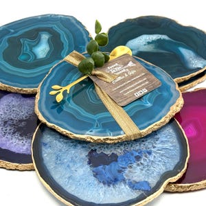 May include: A collection of agate coasters in shades of blue and purple, with gold-painted edges. A gift tag with the text "Found in the Ground Candles & Gifts" is attached with a gold ribbon and decorative sprig.