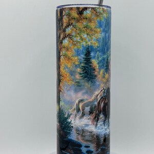 以下が含まれることがあります： 森の川を走る馬のカラフルな画像が描かれたステンレス製のタンブラー。画像は青い空、緑の木々、岩のある茶色の川を表しています。タンブラーにはストローが付いています。