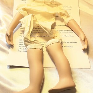May include: A doll kit with a doll's body, arms, and legs, partially assembled. The doll is wearing a cream-colored dress. The kit includes instructions and materials for completing the doll, with text that reads "DOLL IN GREEN COAT".
