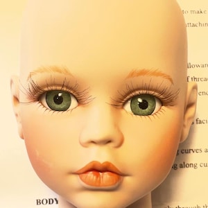 May include: A porcelain doll head with green eyes, long eyelashes, and peach-colored cheeks and lips. The doll head is bald and has a smooth, pale complexion. The word "Jessica" is visible in the background.