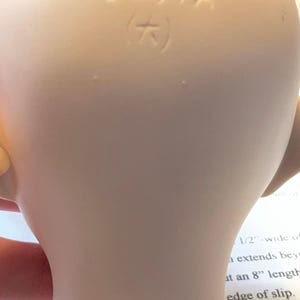 May include: Close-up of a pale, smooth ceramic object with the stamped text "9291A" and a symbol. The object has a curved shape and appears to be part of a larger item. The background includes text.
