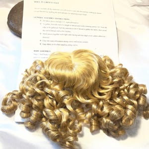 May include: A doll wig with long, golden-blonde curls, resting on a white surface. The wig is the focal point, with a paper instruction sheet titled "Jessica DOLL IN GREEN COAT" visible in the background. The instructions detail assembly steps.