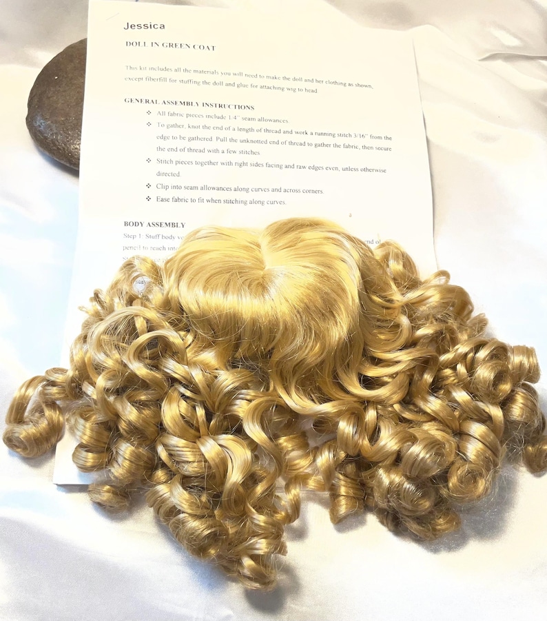 May include: A doll wig with long, golden-blonde curls, resting on a white surface. The wig is the focal point, with a paper instruction sheet titled "Jessica DOLL IN GREEN COAT" visible in the background. The instructions detail assembly steps.