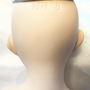 May include: A vintage doll head with a green cap. The doll's head is a pale skin tone, with visible ears and the text "9379 B" on the back of the head. The cap is a muted green color.