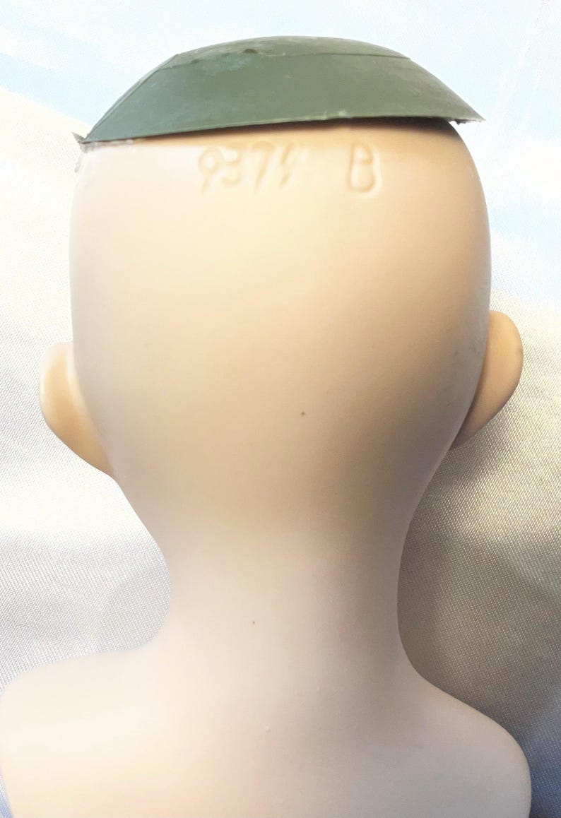 May include: A vintage doll head with a green cap. The doll's head is a pale skin tone, with visible ears and the text "9379 B" on the back of the head. The cap is a muted green color.