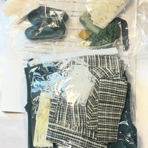 May include: A doll-making kit containing various fabric pieces, lace, and small accessories. The kit includes a green and white plaid fabric, green fabric, and a pair of green doll shoes. Instructions are visible in the background.