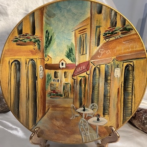 May include: A round decorative plate featuring a hand-painted scene of a European street cafe. The artwork displays yellow buildings, a red awning with the word CAFE, a hanging sign reading CAFE, and two small white tables with chairs on a patio.