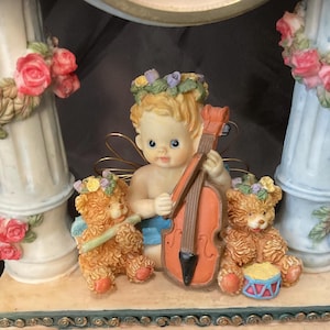 May include: A decorative figurine featuring a cherub playing a violin, flanked by two teddy bears. The cherub and bears are adorned with floral crowns and wings. The figurine is accented with pink roses and decorative columns.