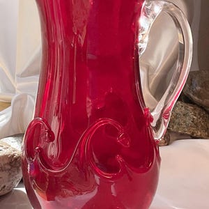 May include: A ruby red glass pitcher with a clear glass handle and base. The pitcher features decorative swirls on the front and a spout for pouring. The base is approximately 3 inches in diameter.