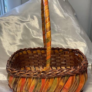 May include: A rectangular woven basket featuring a single upright handle. The basket is constructed from strips of material in shades of brown, orange, and green, with a braided rim detail and a flat base for stability on a flat surface.