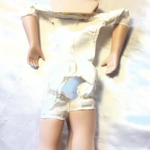 May include: A vintage doll with a porcelain head, arms, and legs. The doll has a painted face with blue eyes, red lips, and rosy cheeks. It is wearing a white satin romper. The doll's hair is missing.