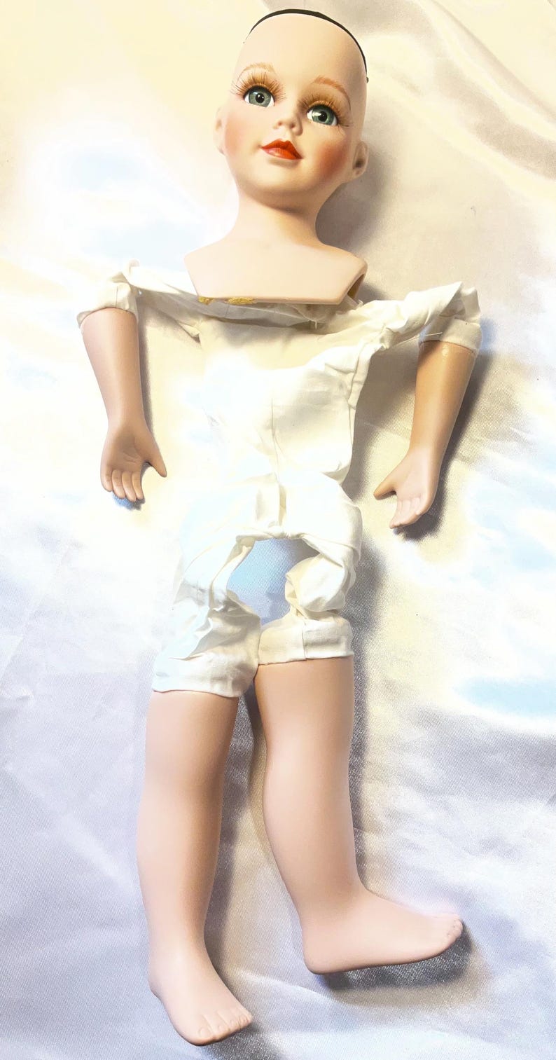 May include: A vintage doll with a porcelain head, arms, and legs. The doll has a painted face with blue eyes, red lips, and rosy cheeks. It is wearing a white satin romper. The doll's hair is missing.