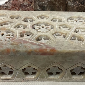 May include: A rectangular, light-colored marble box with intricate carved geometric patterns. The lid features a series of openwork hexagons and starburst designs. The front of the box has star-shaped cutouts. A decorative storage item.