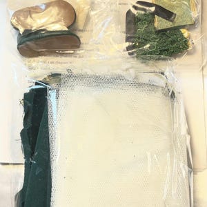 May include: A doll-making kit with various components visible through clear packaging. Includes small doll shoes, green fabric, a white mesh, and other accessories. The kit appears to contain materials for creating a doll.