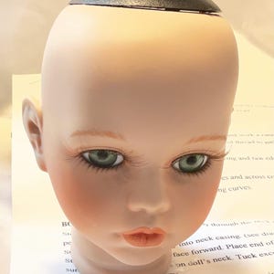 May include: A doll head with a pale complexion, green eyes, and long eyelashes. The doll head has a black cap on top and is resting on a piece of paper with instructions.