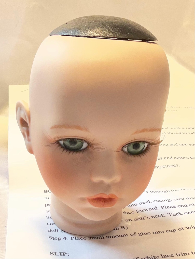 May include: A doll head with a pale complexion, green eyes, and long eyelashes. The doll head has a black cap on top and is resting on a piece of paper with instructions.