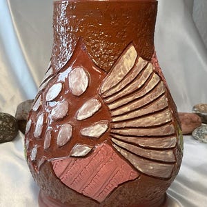 May include: A handcrafted ceramic vase with a textured, brown exterior. The vase features raised, decorative elements in shades of pink, white, and brown. The base is a solid, round shape. The vase is approximately 8 inches tall.