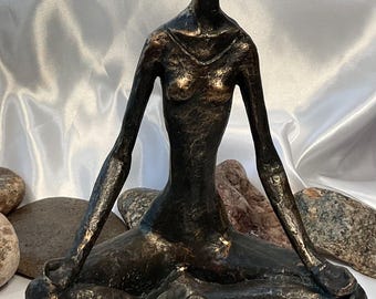 May include: Bronze-toned sculpture of a seated figure in a yoga pose. The figure has elongated limbs and is seated on a dark, textured base. The sculpture is set against a backdrop of white fabric and stones.