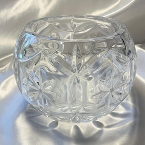 May include: A clear, spherical glass bowl featuring a prominent starburst or snowflake-like cut pattern across its surface. The bowl has a wide, circular opening at the top and a thick, transparent glass construction with reflective facets.