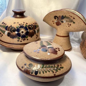 May include: A collection of handcrafted pottery pieces with floral designs. The set includes a domed container, a lidded jar, and a napkin holder. The pottery features a tan base color with brown, blue, green, and white painted accents.