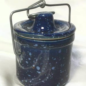 May include: A cylindrical ceramic crock with a dark blue glaze and light blue speckled pattern. The container features a matching lid with a central knob and a hinged metal wire handle that arches over the top for carrying or hanging purposes.