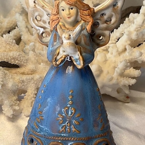 May include: A ceramic angel figurine with a blue dress and gold accents. The angel has light brown hair and holds a small white rabbit. The wings are beige with decorative cutouts. The base of the figurine has a decorative pattern.