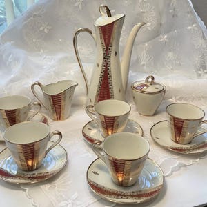 May include: A vintage tea set featuring a tall coffee pot, creamer, sugar bowl, and six teacups with saucers. The set has a cream base with gold and red accents. The coffee pot is approximately 10 inches tall. The set is arranged on a white tablecloth.