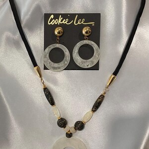 May include: A white and gold necklace with a large white circle pendant and black and white beads. The necklace is on a black cord. The image also shows a pair of white circle earrings with gold accents. The earrings are on a black card with the text "Cookee Lee" printed on it.
