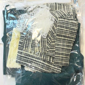 May include: A sealed plastic bag containing various fabric pieces for a craft project. The fabrics include a black and white plaid, dark green, and a cream-colored lace trim. Also included is a yellow fabric piece and other small accessories.