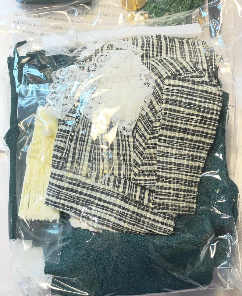 May include: A sealed plastic bag containing various fabric pieces for a craft project. The fabrics include a black and white plaid, dark green, and a cream-colored lace trim. Also included is a yellow fabric piece and other small accessories.