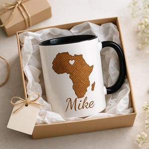 May include: White ceramic mug with a black handle and rim, featuring a brown Africa map design and the name "Mike". The mug is presented in a gift box with tissue paper, a gift tag, and a decorative bow. A thoughtful gift idea.