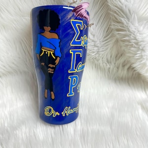 May include: A blue and gold tumbler with a black and white illustration of a woman wearing a blue shirt and black pants. The tumbler has the letters "ΣΓΡ" and the text "Dr. Hampton" on it.