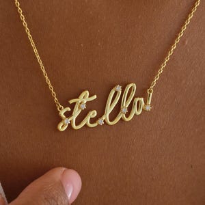 May include: A gold-colored necklace with the name "Stella" in cursive script. The necklace has small, sparkling accents on the letters. The necklace is worn around the neck.