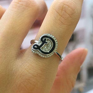 以下が含まれることがあります： 黒いエナメルで「D」の文字が施されたシルバーリング。小さな透明な石のハローで囲まれています。指にはめられ、エレガントなデザインと職人技が際立っています。リングのバンドは細く、磨かれています。