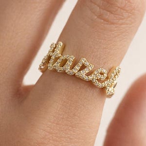 Peut inclure: Une bague dorée avec le nom "Hazel" écrit en cursive. La bague est ornée de petits diamants transparents. Le bijou est porté au doigt.