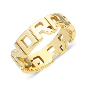 Puede incluir: Un anillo dorado con la palabra "DREAM" escrita en letras de bloque. El anillo tiene un acabado pulido y un diseño geométrico moderno. Las letras están conectadas, formando una banda continua. Hecho de metal brillante.